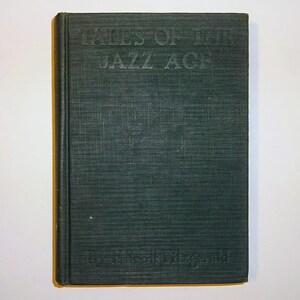 May include: A vintage hardcover book titled "Tales of the Jazz Age" by F. Scott Fitzgerald. The book has a dark green cover with embossed lettering. The book is closed and set against a white background.
