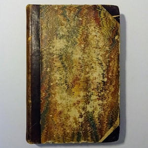 May include: A vintage book with a brown leather spine and a marbled cover with faded colors. The cover is worn and has some stains.