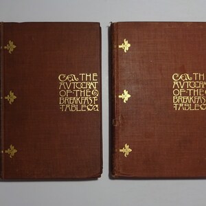 May include: Two brown hardback books with gold lettering on the spine and front cover. The title of the book is "The Autocrat of the Breakfast Table".