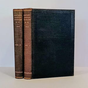 May include: Two antique books, "History of the Town of Durham, N.H. 1913," are displayed. Volume II is on the left, Volume I is in the middle, and a black book is on the right. The spines are brown and the text is in gold.
