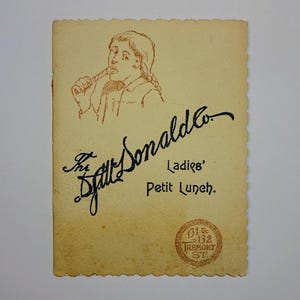 May include: A vintage menu card for The Doll Donald Co. Ladies' Petit Lunch. The card features a brown line drawing of a woman with a braid, holding a piece of food. The card has a brown border and a brown seal with the text "81 & 132 TREMONT ST."