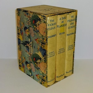 May include: A set of three vintage children's books in a slipcase. The books are titled "The Cuckoo Clock", "A Dog of Flanders", and "The Princess and Curdie". The slipcase is decorated with a colorful illustration of children playing in a garden.