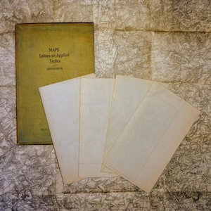 May include: A vintage book titled "Maps Letters on Applied Tactics" by Greenhill, with a yellow cover and a stack of blank white papers on top of it.