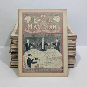 Puede incluir: Una revista antigua titulada The Eagle Magician de noviembre de 1915. La portada presenta una ilustraci&oacute;n gris y roja de magos realizando trucos, incluido un conejo saliendo de un sombrero. El texto incluye Volume, Number y Price 10-Cents.