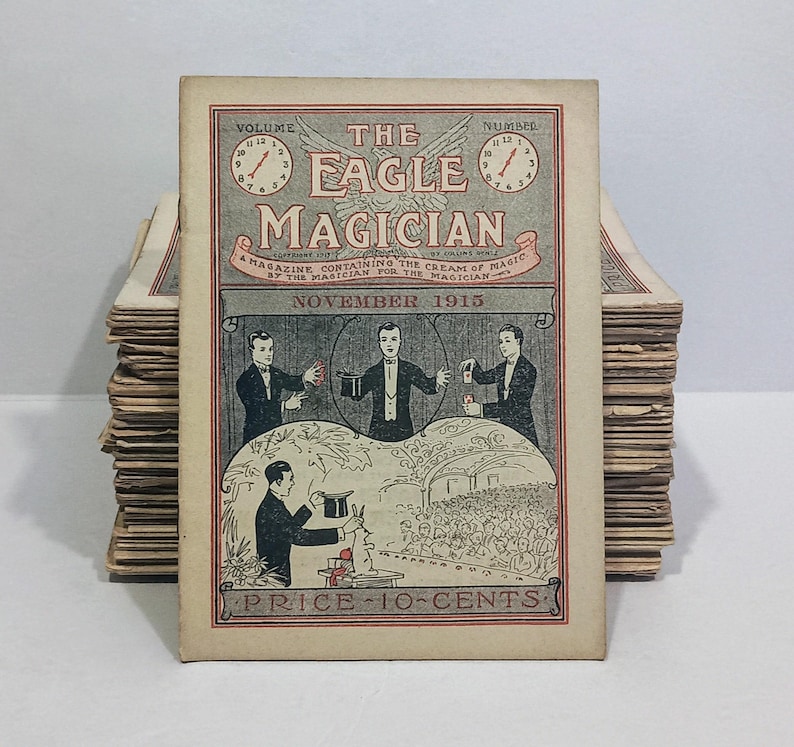Puede incluir: Una revista antigua titulada The Eagle Magician de noviembre de 1915. La portada presenta una ilustraci&oacute;n gris y roja de magos realizando trucos, incluido un conejo saliendo de un sombrero. El texto incluye Volume, Number y Price 10-Cents.