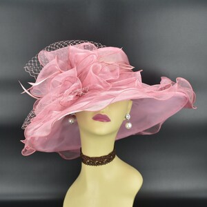 May include: A pink, wide-brimmed hat with a layered, ruffled design and a delicate veil. The hat is displayed on a mannequin head, showcasing its elegant shape and texture. Pearl earrings and a brown choker complete the look.