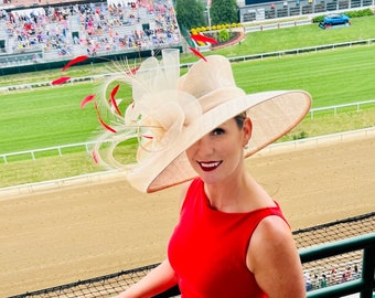 May include: A woman wearing a large, wide-brimmed, cream-colored hat with red feathers and a red dress. She is standing on a balcony overlooking a racetrack.