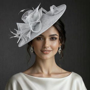May include: A woman wearing a light gray hat with a decorative floral accent and feathers. The hat has a wide brim and is paired with a white satin top. The background is a dark gray.