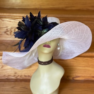 May include: A white wide-brimmed hat with a large navy blue and green feather embellishment. The hat is on a mannequin head.