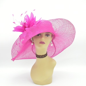 May include: A large, bright pink, wide-brimmed hat with a feather accent. The hat is made of a straw-like material and has a slightly curved brim.
