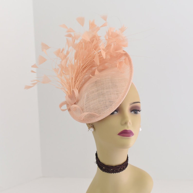May include: A peach-colored fascinator hat with a large, round brim and a feather plume. The hat is made of a delicate, woven material and is perfect for a special occasion.