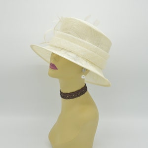 Op de afbeelding: Een cr&egrave;mekleurige hoed met een brede rand en een gestructureerde kroon. De hoed heeft een decoratieve band en is versierd met delicate veren. Een mannequinhoofd toont de hoed, samen met een bruine kanten choker en een parel oorbellen.
