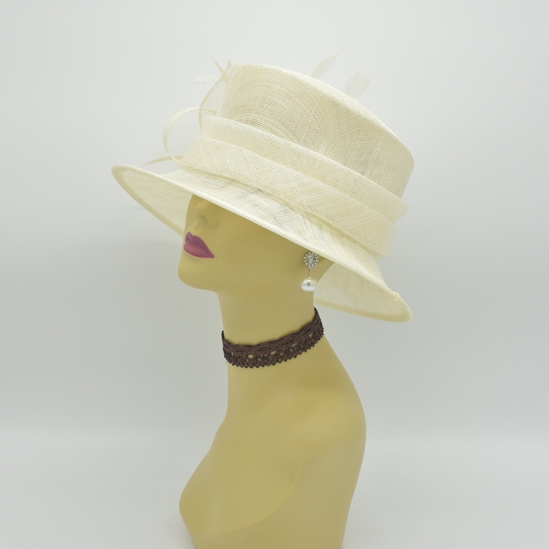 Op de afbeelding: Een cr&egrave;mekleurige hoed met een brede rand en een gestructureerde kroon. De hoed heeft een decoratieve band en is versierd met delicate veren. Een mannequinhoofd toont de hoed, samen met een bruine kanten choker en een parel oorbellen.