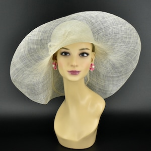 May include: A cream-coloured, wide-brimmed hat with a textured weave, displayed on a mannequin head. The hat has a rounded crown and a large, floppy brim. The mannequin wears pink flower earrings with a pearl centre. The background is black.