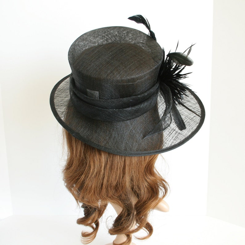 May include: A black hat with a wide brim and a black feather trim. The hat is made of a straw-like material.