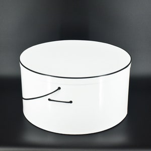 May include: White round hat box with a black trim around the top edge. The box has a black string handle.