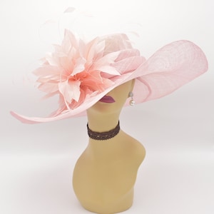 May include: A wide-brimmed hat in pink with a large pink flower on the side. The hat is made from a straw-like material and has a wide brim that curves down at the sides.