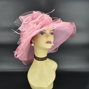 May include: A light pink, wide-brimmed hat with a decorative floral accent and a delicate veil. The hat is displayed on a mannequin head, showcasing its elegant design. The hat is suitable for special occasions.