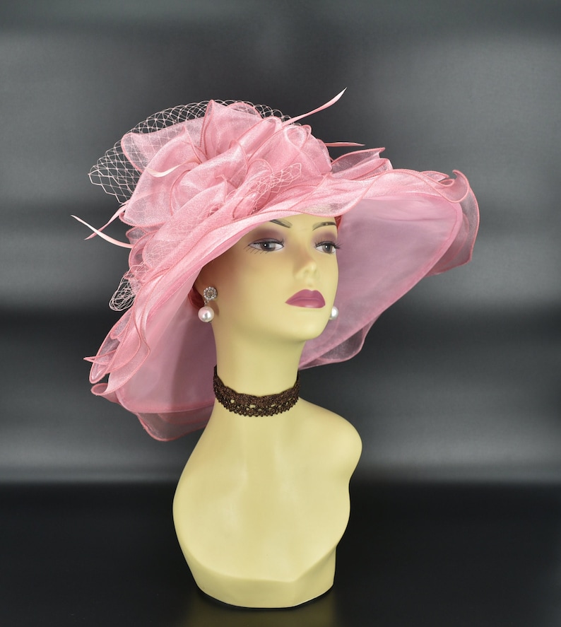 May include: A light pink, wide-brimmed hat with a decorative floral accent and a delicate veil. The hat is displayed on a mannequin head, showcasing its elegant design. The hat is suitable for special occasions.