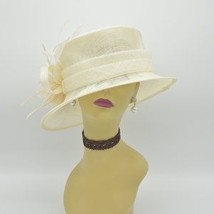 Op de afbeelding: Cr&egrave;mekleurige hoed met een brede rand en een decoratieve band. De hoed heeft een strik en veren aan de zijkant. De mannequin draagt pareloorbellen en een bruine kanten choker. Een formele accessoire.