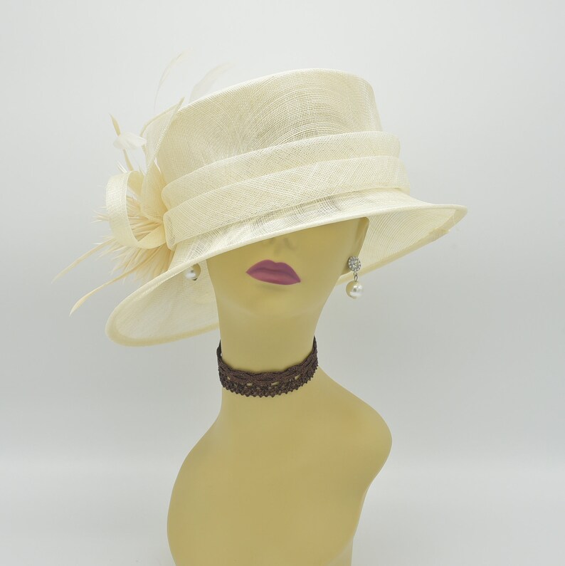 Op de afbeelding: Cr&egrave;mekleurige hoed met een brede rand en een decoratieve band. De hoed heeft een strik en veren aan de zijkant. De mannequin draagt pareloorbellen en een bruine kanten choker. Een formele accessoire.