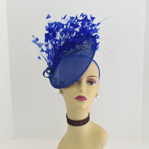 May include: A royal blue fascinator hat with a large, curled feather plume. The hat is made of a woven fabric and has a wide brim.