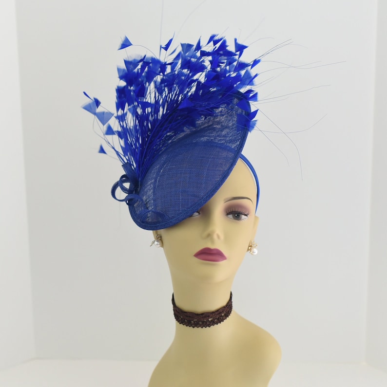 May include: A royal blue fascinator hat with a large, curled feather plume. The hat is made of a woven fabric and has a wide brim.