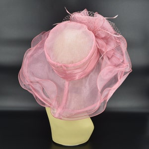 May include: A pink formal hat with a wide brim and a rounded crown. The hat features a sheer, ruffled fabric and is adorned with a decorative bow and netting. The hat is displayed on a mannequin head.