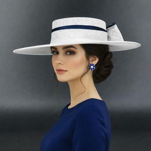 Puede incluir: Un sombrero blanco de ala ancha con copa plana y una cinta azul oscuro que rodea la base, con un lazo estructurado en el lateral. El sombrero está confeccionado en un material trenzado y se lleva con una parte superior azul oscuro y pendientes de flores.