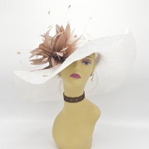 May include: A white wide-brimmed hat with a brown feather accent. The hat is made of a straw-like material and has a large brim that curves down on the sides. The feather accent is a large, fluffy plume that is attached to the hat's crown.