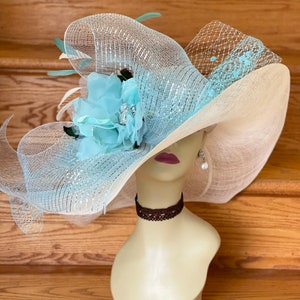Peut inclure: Un grand chapeau à larges bords avec un design bleu clair et blanc. Le chapeau est décoré d'une grande fleur bleu clair et d'une garniture argentée. Le chapeau est posé sur la tête d'un mannequin.
