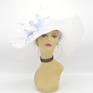 May include: A white wide-brimmed hat with a blue and white flower accent. The hat is made of a straw-like material and has a wide brim that curves down on the sides. The flower is made of fabric and has a light blue colour with white accents.