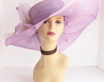 May include: A lavender-colored wide-brimmed hat with a large bow and feather detail. The hat is made of a soft, delicate material and has a wide, floppy brim.