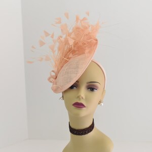 May include: A peach-colored fascinator hat with a large feather plume. The hat is made of a soft, delicate material and has a wide brim.