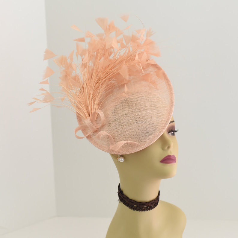 May include: A peach-colored hat with a large, round brim and a feather accent. The hat is made of a straw-like material and has a delicate, feminine design.