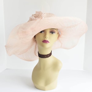 May include: A large, wide-brimmed hat made of a delicate, pale pink fabric. The hat has a ruffled trim and a small, white flower accent. The hat is worn on a mannequin with a brown choker.