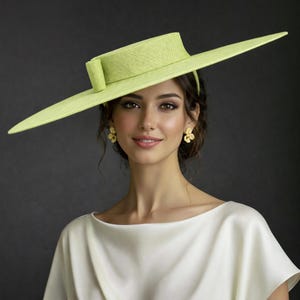 May include: A light green wide-brimmed hat with a rectangular crown and a decorative bow. The hat is made of woven material. The woman is wearing a white top and gold flower earrings. The background is dark.