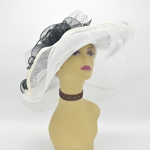 May include: A large, white brimmed hat with a black and cream-coloured decorative feature. The hat has a sheer veil and is displayed on a mannequin head. This is a formal accessory.