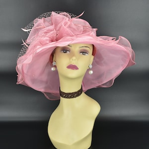 May include: A light pink, wide-brimmed hat with a sheer, ruffled design and a delicate veil. The hat is displayed on a mannequin head, paired with pearl earrings and a brown lace choker. The hat's brim measures approximately 16 inches in diameter.