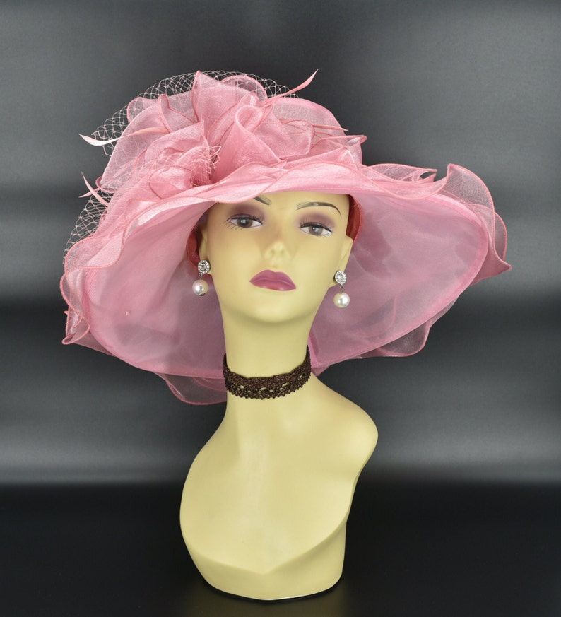 May include: A light pink, wide-brimmed hat with a sheer, ruffled design and a delicate veil. The hat is displayed on a mannequin head, paired with pearl earrings and a brown lace choker. The hat's brim measures approximately 16 inches in diameter.