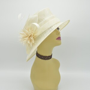 Op de afbeelding: Een cr&egrave;mekleurige hoed met een brede rand en een gelaagde kroon. De hoed is versierd met een beige bloem en veren aan de zijkant. Een bruine kanten choker zit om de nek van de mannequin.
