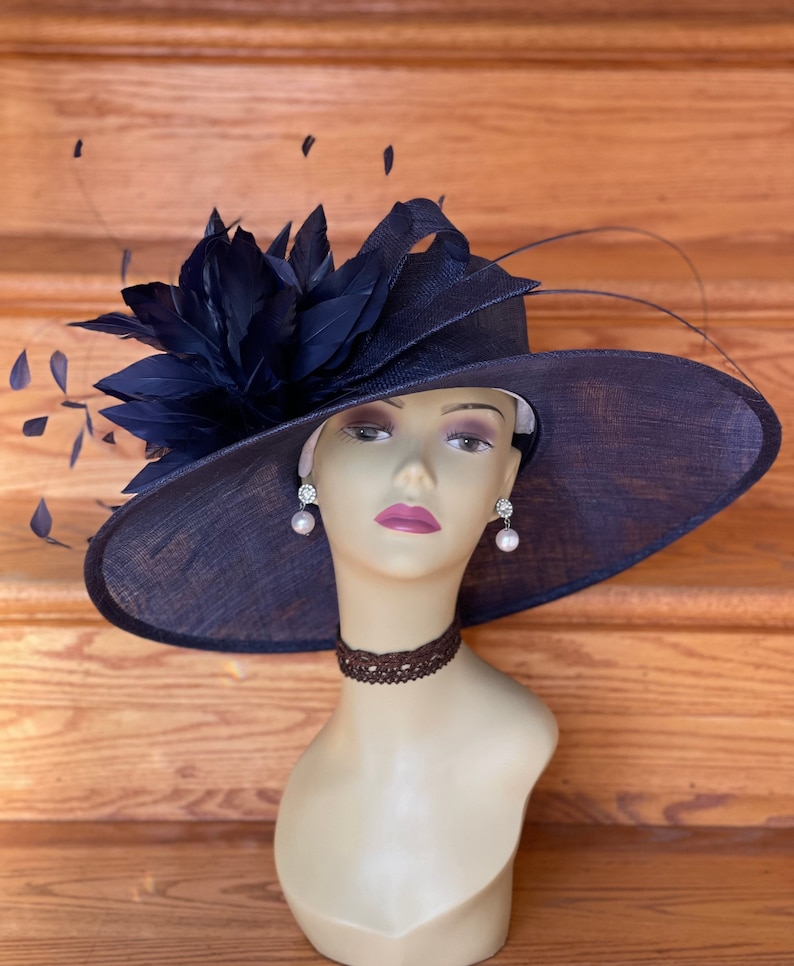 K&ouml;nnte beinhalten: Ein breitkrempiger, marineblauer Hut mit einem gro&szlig;en, marineblauen Federschmuck. Der Hut ist aus stroh&auml;hnlichem Material gefertigt und hat eine breite Krempe, die an den Seiten nach unten gebogen ist. Der Hut sitzt auf dem Kopf einer Schaufensterpuppe.