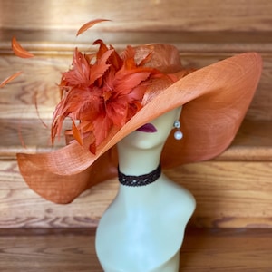 May include: A large, wide-brimmed hat in a burnt orange color with a matching feather embellishment. The hat is made of a straw-like material and is sitting on a mannequin.