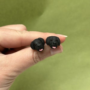 K&ouml;nnte beinhalten: Ein Paar schwarze Labrador Retriever Ohrstecker. Jeder Ohrring zeigt eine detaillierte Miniatur-Skulptur eines Hundekopfes mit rosa Zunge. Die Ohrringe sind vor einem weichen, gr&uuml;nen Hintergrund platziert.