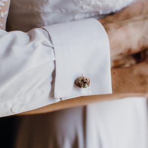 Puede incluir: Primer plano de un puño de camisa blanca con un gemelo decorativo. El gemelo tiene un diseño pequeño, redondo y texturizado con tonos marrones y verdes. La imagen sugiere una boda o una ocasión formal.