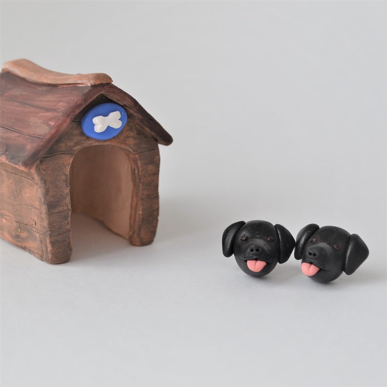 K&ouml;nnte beinhalten: Ein braunes Hundeh&auml;uschen mit einem blau-wei&szlig;en Knochenemblem, neben zwei schwarzen Hundekopfskulpturen mit rosa Zungen. Das Hundeh&auml;uschen hat ein holzartiges Aussehen.