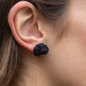 Könnte beinhalten: Ein Paar schwarze Ohrstecker mit einem strukturierten 3D-Design. Die Ohrringe werden im Ohrläppchen einer Person getragen.