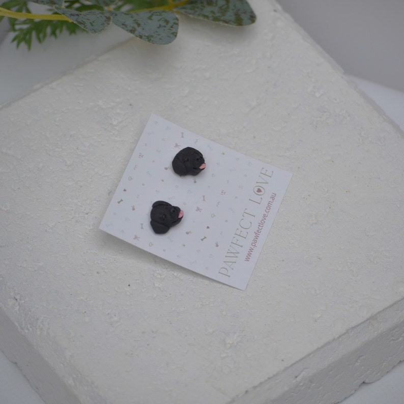 K&ouml;nnte beinhalten: Schwarze Mops-Ohrstecker mit rosa Zungen, pr&auml;sentiert auf einer wei&szlig;en Karte mit dem Text "PAWFECT LOVE". Die Ohrringe befinden sich auf einer strukturierten wei&szlig;en Oberfl&auml;che. Ein niedliches und einzigartiges Accessoire.