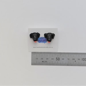 K&ouml;nnte beinhalten: Ein Paar schwarze Ohrstecker in Form von Hundek&ouml;pfen mit rosa Zungen, pr&auml;sentiert auf einer wei&szlig;en Karte. Ein Metalllineal ist darunter platziert. Die Ohrringe sind handgefertigt.