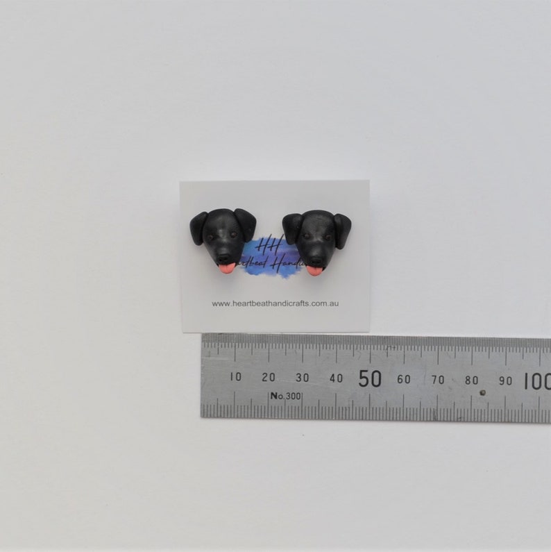 K&ouml;nnte beinhalten: Ein Paar schwarze Ohrstecker in Form von Hundek&ouml;pfen mit rosa Zungen, pr&auml;sentiert auf einer wei&szlig;en Karte. Ein Metalllineal ist darunter platziert. Die Ohrringe sind handgefertigt.